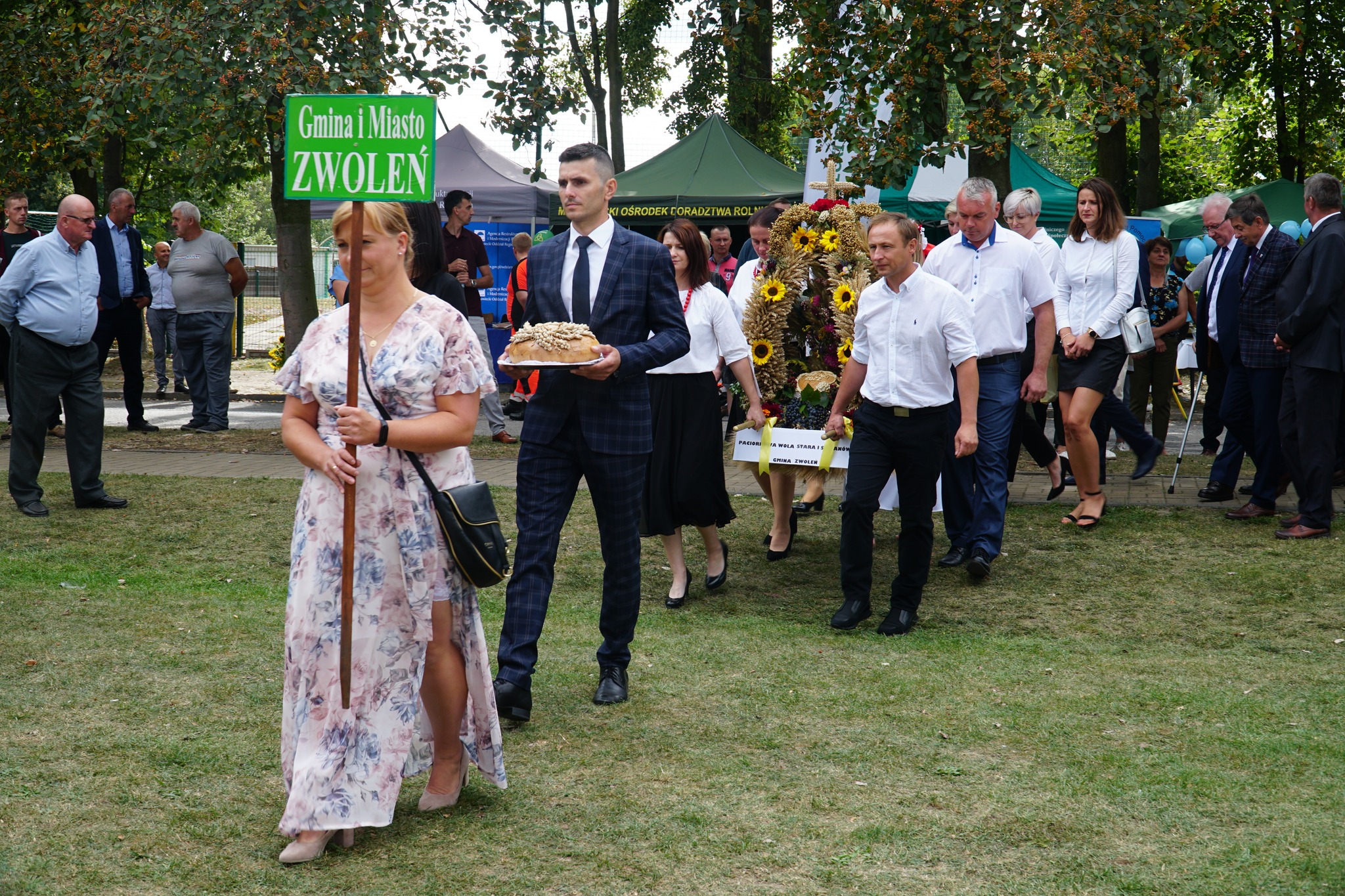 Zdjęcie przedstawia grupę osób. Pierwsza kobieta trzyma tabliczkę z napisem Zwoleń, mężczyzna za nią trzyma w rękach chleb, następnie cztery osoby niosą wieniec dożynkowy