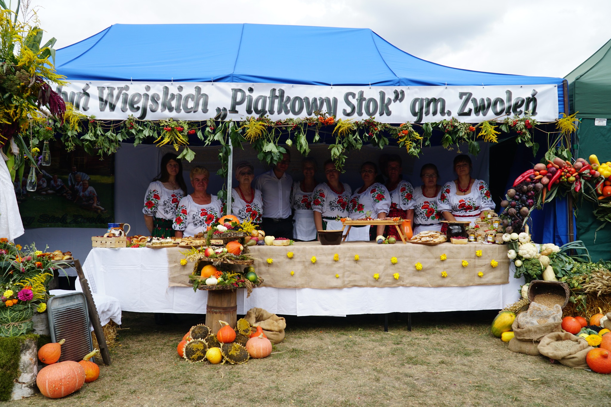 Zdjęcie przedstawia stoisko dożynkowe przygotowana przez Piątkowy Stok. Na drugim planie grupa osób ubrana w folklorystyczne stroje.