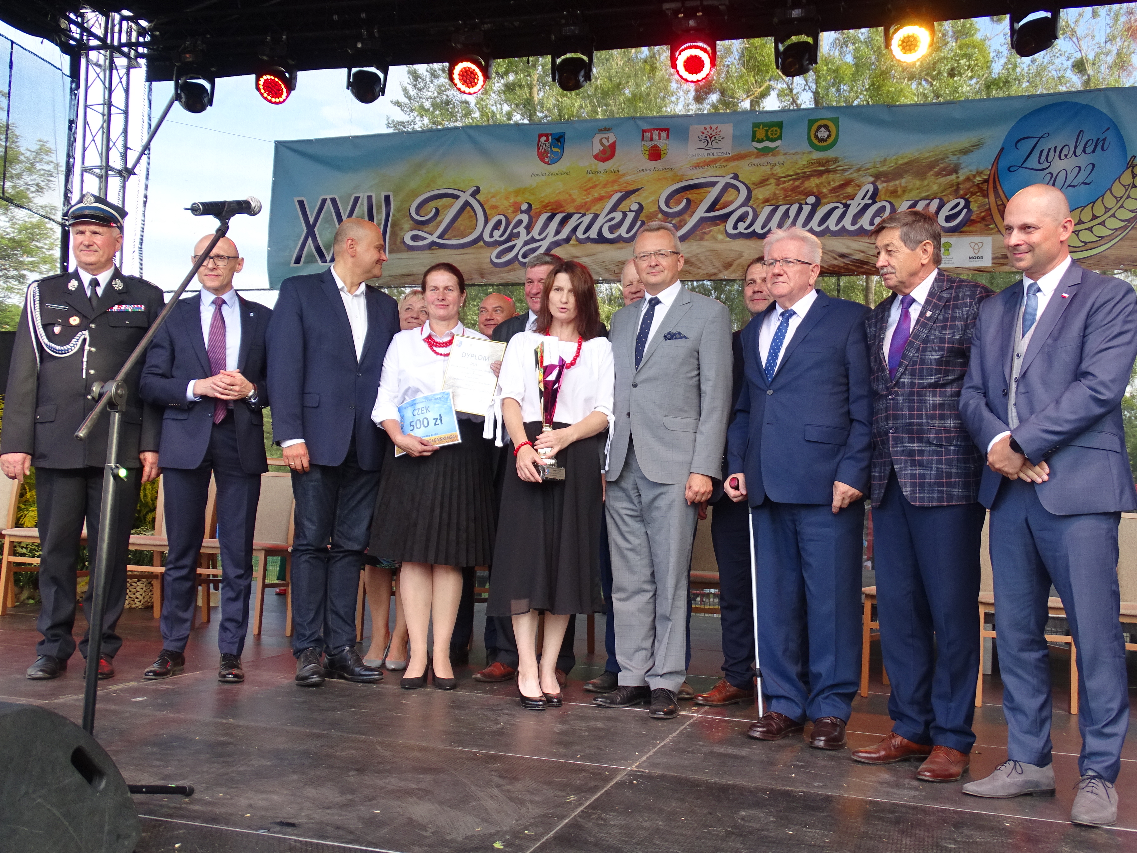 Zdjęcie przedstawia grupę osób stojących przodem na scenie. Jedna z kobiet trzyma symboliczny czek. Kobieta znajdująca się  obok niej trzyma pamiątkowy puchar