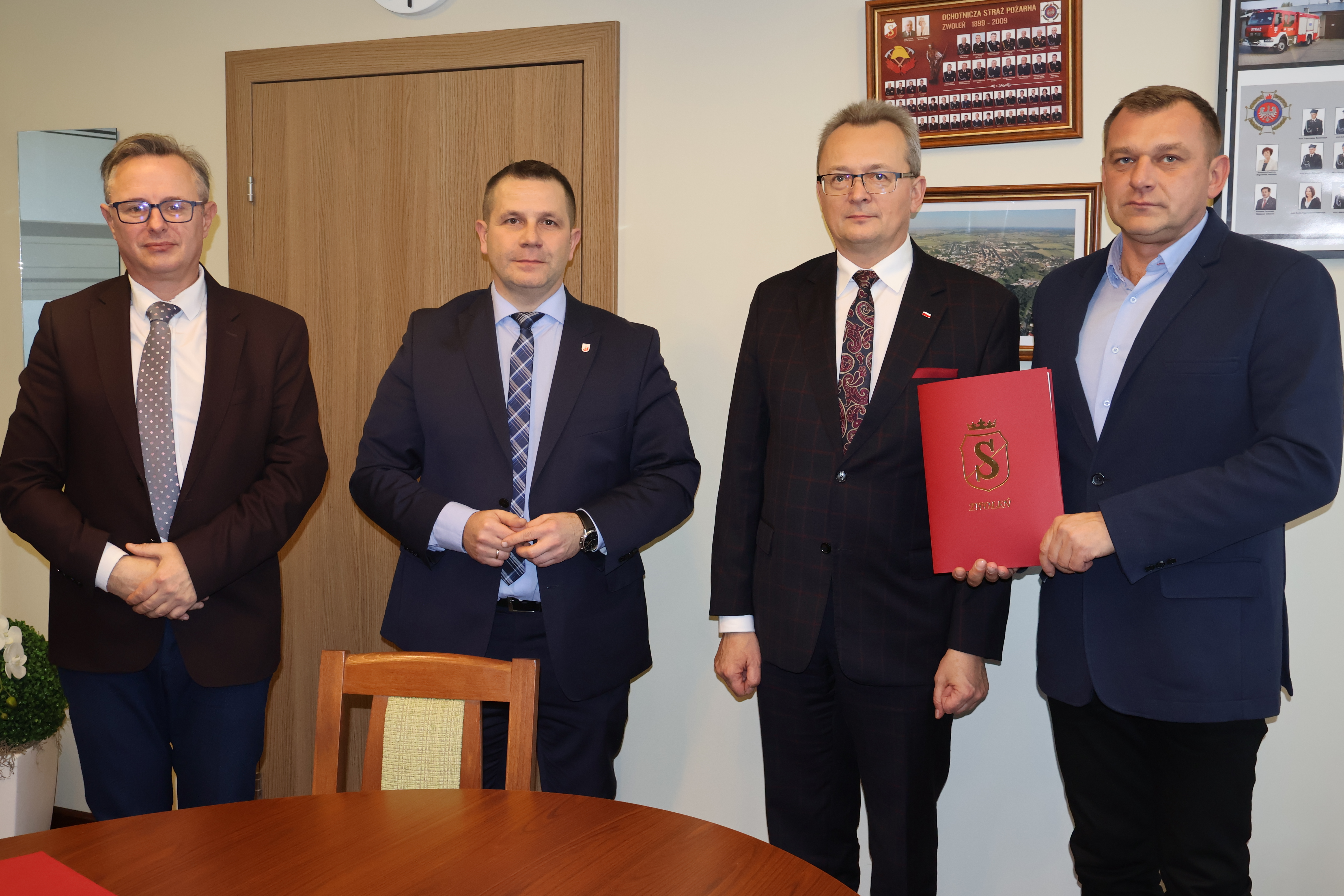 Pamiątkowa fotografia tuż po podpisaniu umowy. Na zdjęciu znajduje się 4 mężczyzn. Jeden z nich trzyma czerwoną teczkę. 