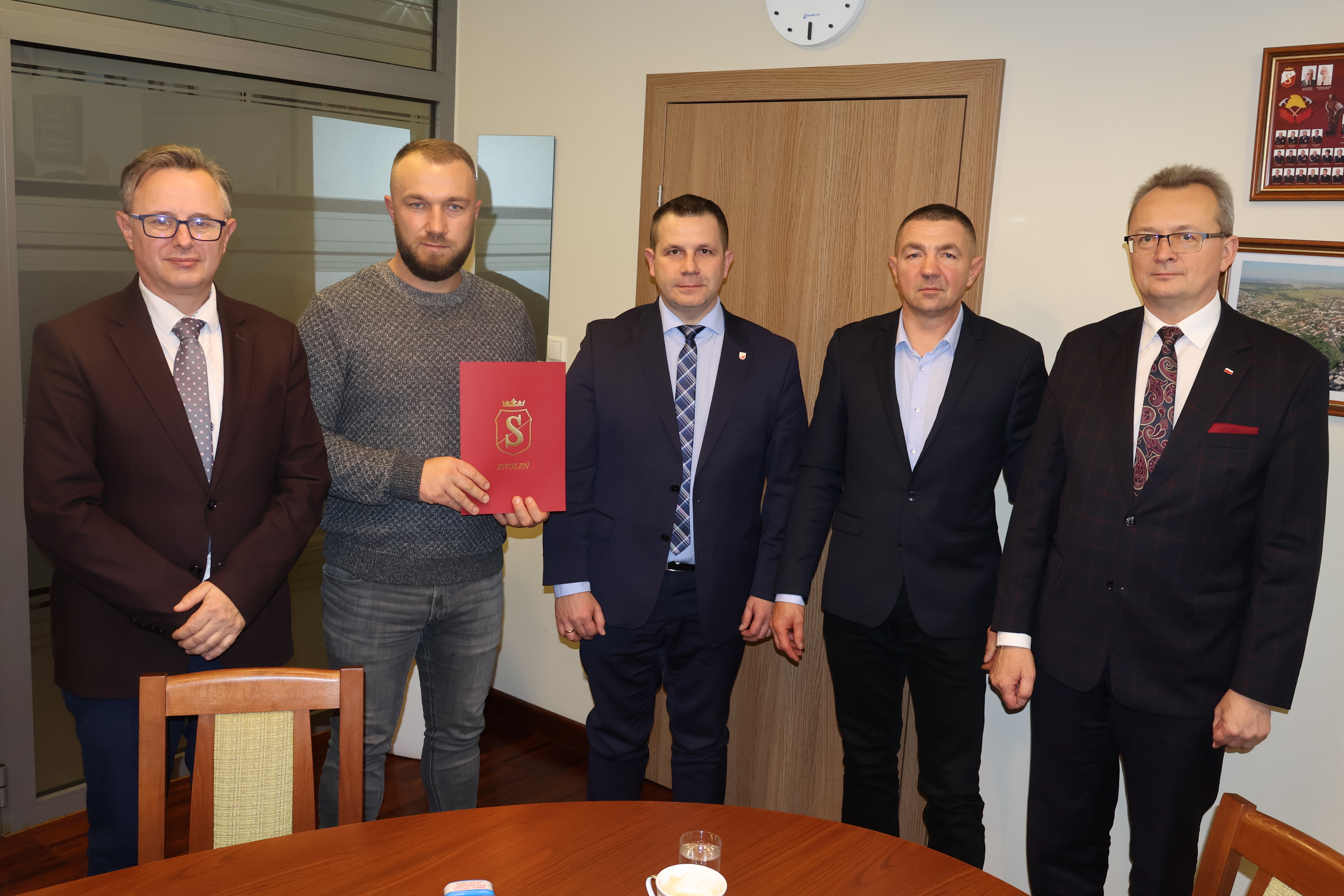 Pamiątkowa fotografia. Na zdjęciu 5 mężczyzn, 1 z nich trzyma czerwoną teczkę 
