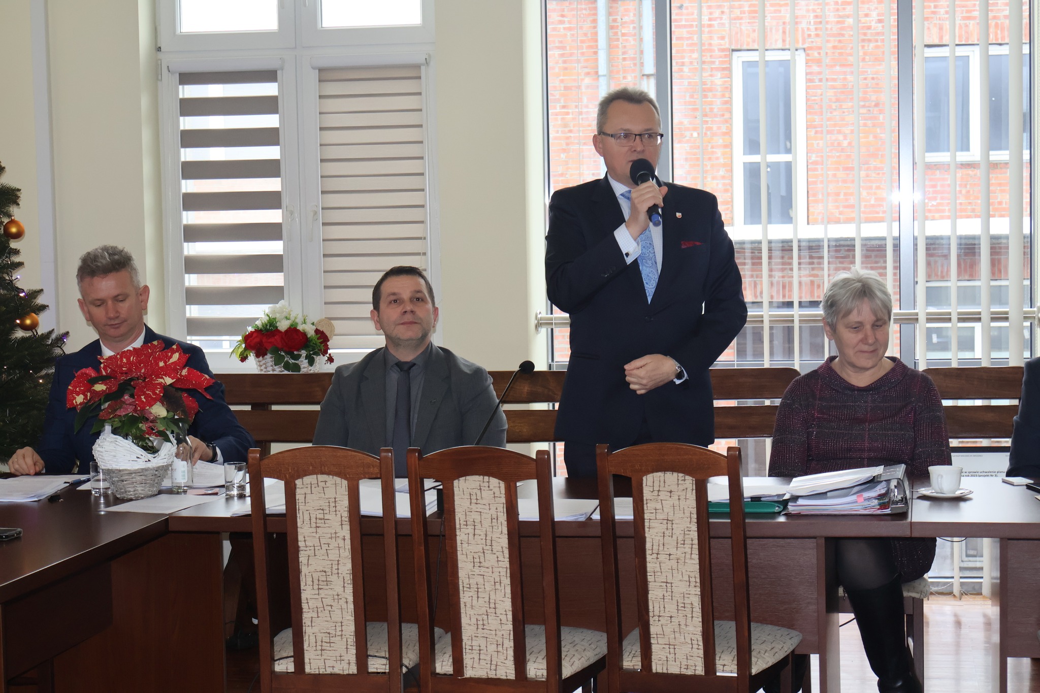 Na zdjęciu znajdują się cztery osoby, jedna z nich stoi i przemawia. 