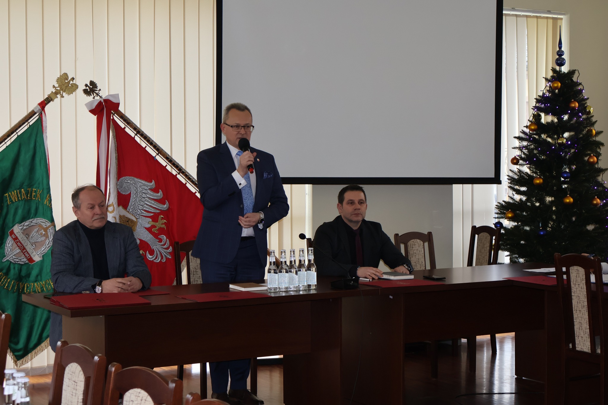 Na zdjęciu jest trzech mężczyzn, jeden z nich stoi i przemawia do mikrofonu. 