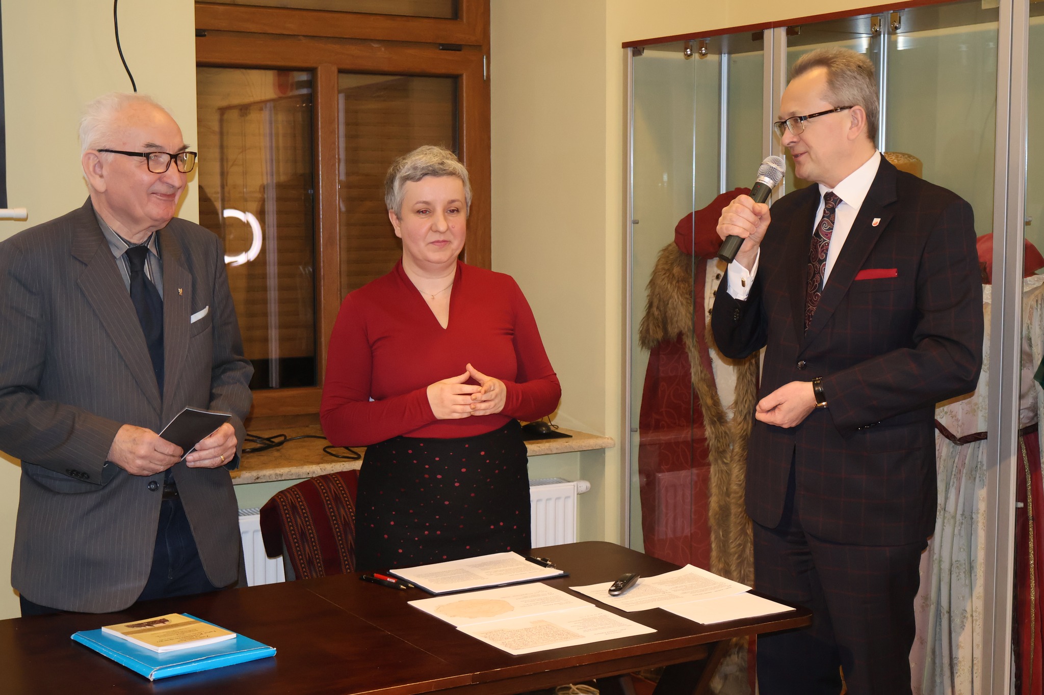 Na zdjęciu znajdują się trzy osoby: dwóch mężczyzn i jedna kobieta. Wszyscy stoją, jden z mężczyzn trzyma w ręku mikrofon 