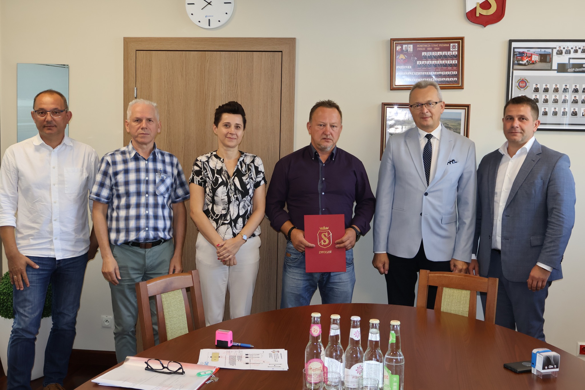 Na zdjeciu znajduje się 6 osób, w tym jedna kobieta. Wszyscy stoją, jeden z mężczyzn trzyma czerwoną teczkę.  